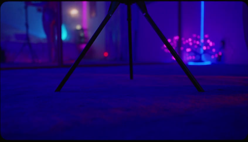 Woman grabbing a tripod and setting it up in front of the camera in a room with black and purple lights