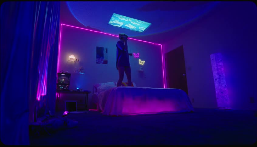 Woman bounces on bed while dancing in bedroom lit by blue and purple colored lighting