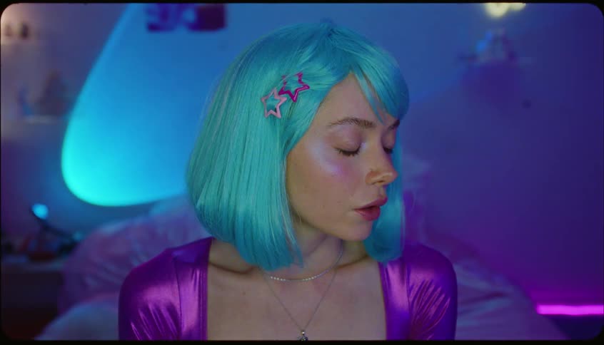 Young lady on lightblue wig smiles while seated on the bed indoor