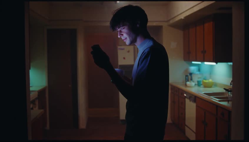 Man sipping from a cup and using his phone in the kitchen at night