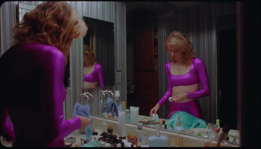 Young woman dressed in purple in a dressing room indoors removing a teal wig and wiping her face with a tissue to remove make up
