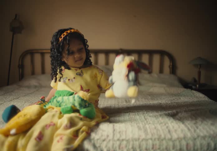 Girl wearing a yellow dress sitting down on a bed indoors playing with stuffed animals