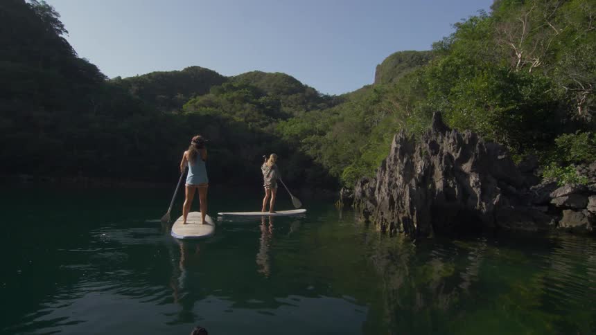 Two women standing on paddle boards and traveling down a calm body of water in the mountains