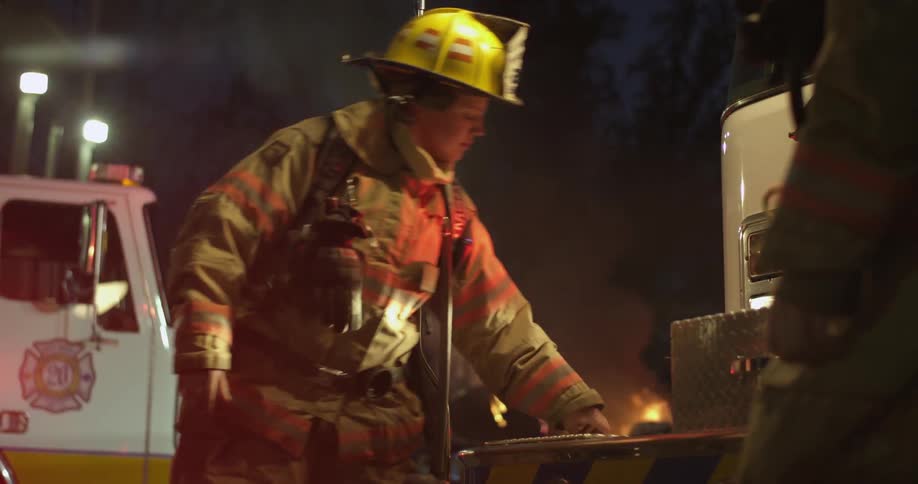 A firefighter walks over to a fire truck and opens a compartment at night
