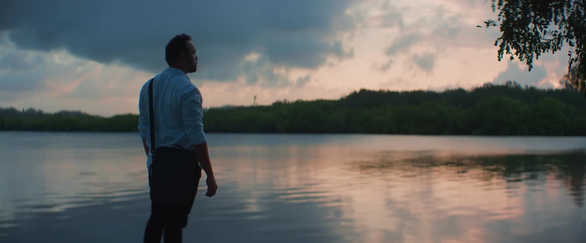 A man stands in front of a river with water reflecting the sky and is admiring the environment