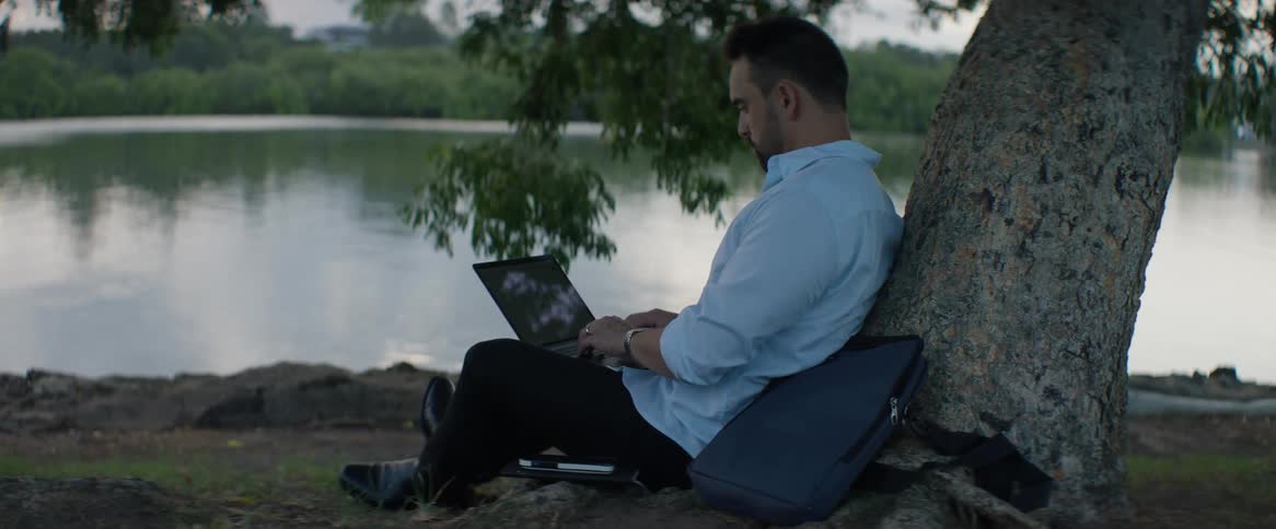 Man leans against tree on lake shore while typing on laptop