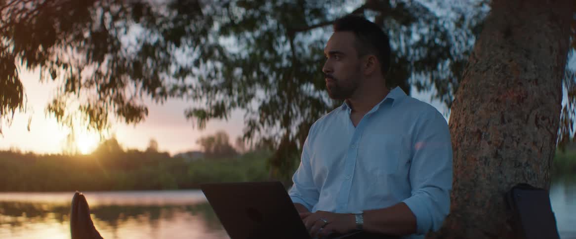 Man sits on shore near tree and types on laptop as sun sets