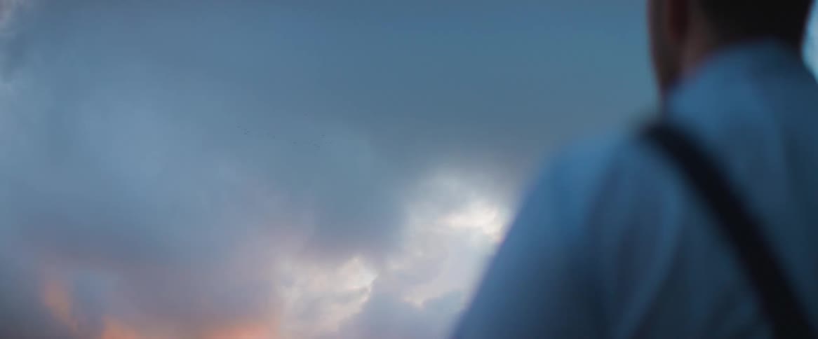 A man wearing a backpack is turning his back and looking at the sky which is the orange color of a sunset