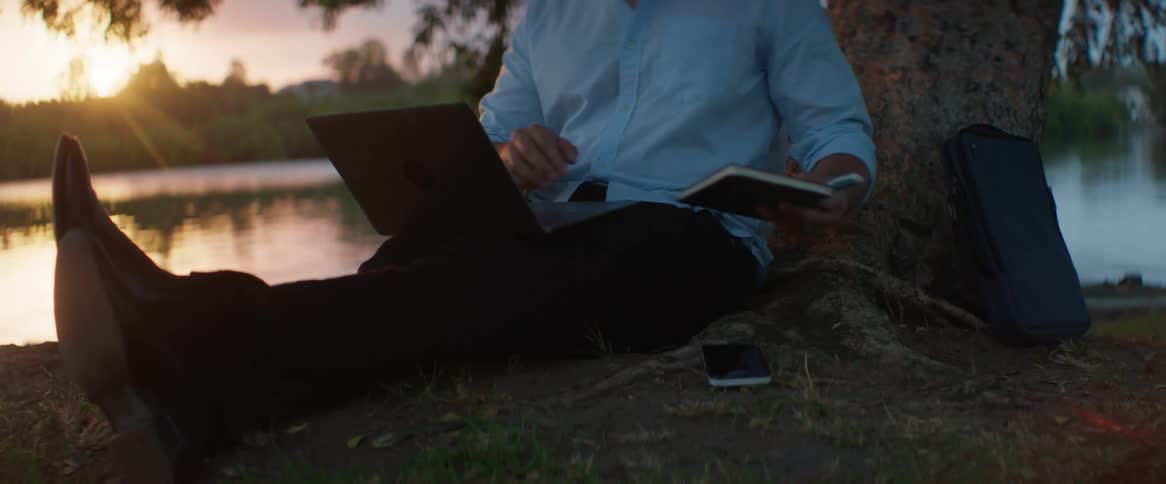 A man sitting beneath a tree with his laptop and picking up a book