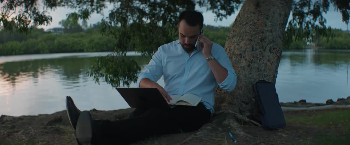 Man sitting under a tree talking on the phone and reading a book at the same time