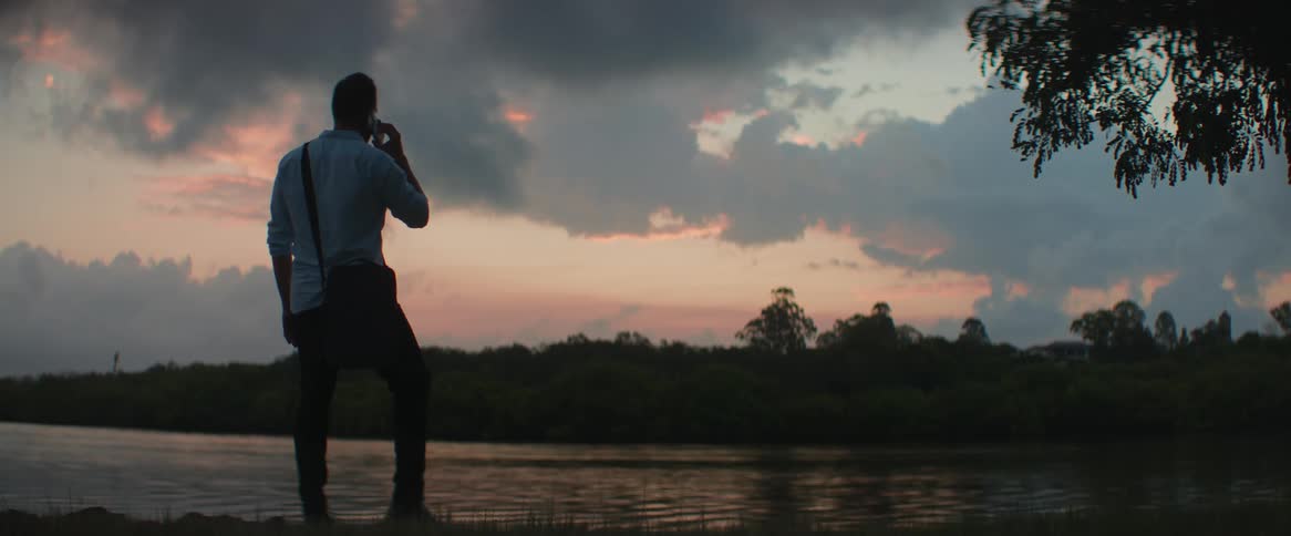 A man stands near a river while making a call at dusk