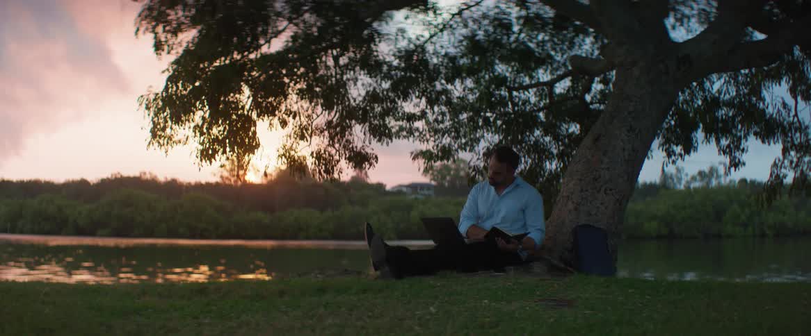Man sits under a tree at dusk writing on a book and a laptop on his lap