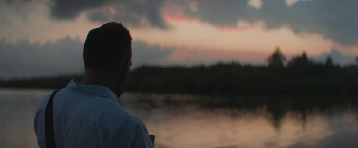 A man talking on his cell phone and then taking a photo of the lake in front of him with the dawn sky