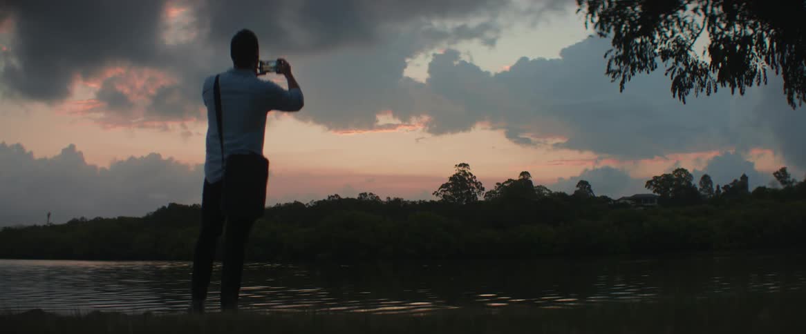 Man standing next to a river takes a picture of the sunset