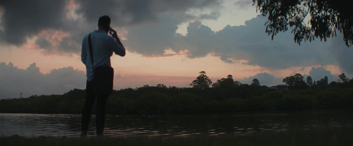 A man is standing on a riverbank talking on his cell phone