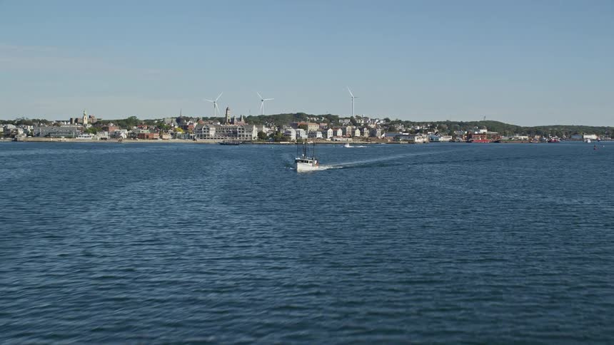 A big ocean with boats passing by and behind it there is a residential area on the coast and windmills
