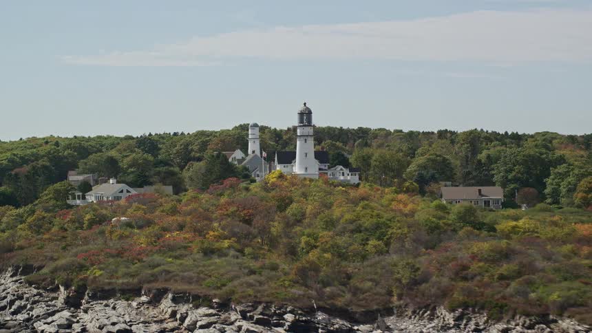 A white lighthouse in a land with some houses facing the sea with several rocks on the coast