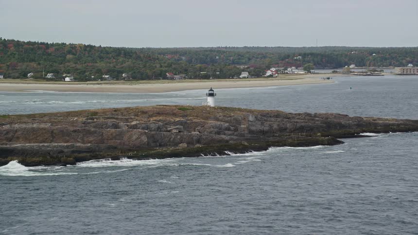 An ilkah with a small white lighthouse in the middle of the sea and behind a beach