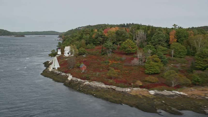 Waves crash against shore near lighthouse and house on forested peninsula during the autumn