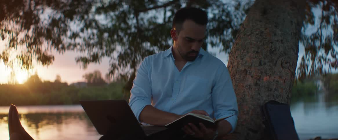 Portrait of a man sitting under a tree writing in his notebook with a lake in the background and the morning sun
