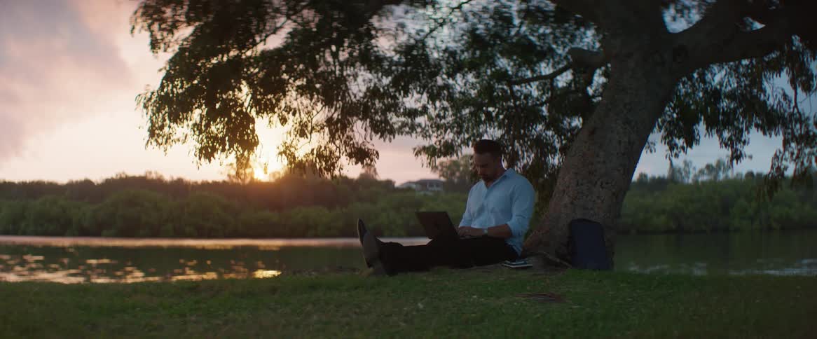 Man sitting under a tree writing on his laptop next to a lake