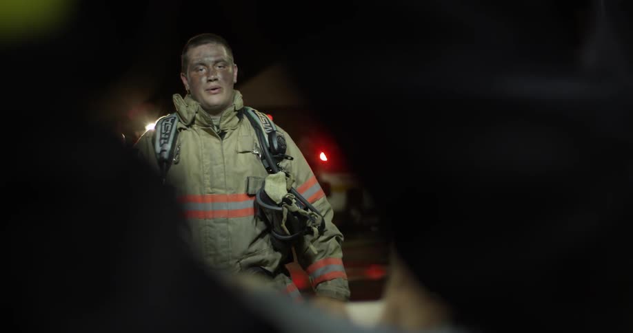 A firefighter standing in front of a fire engine and fire looking at a person