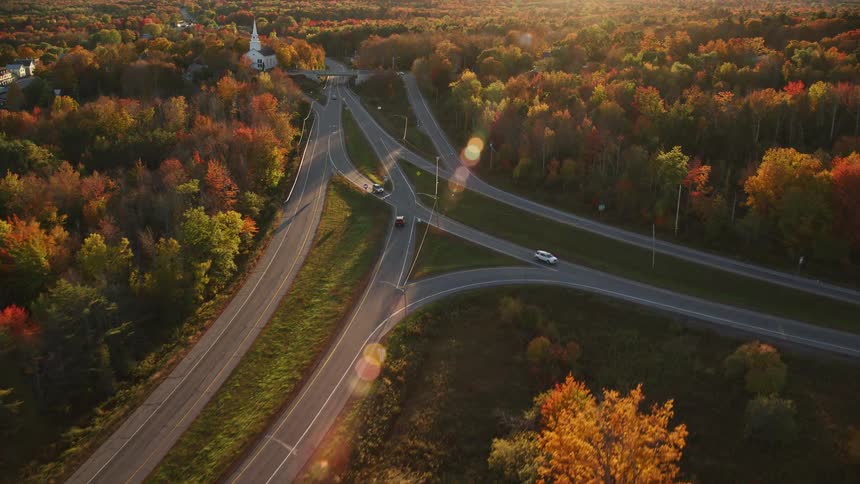 A red truck travels with light traffic on a road surrounded by trees in bright fall colors into a residential area along the coast