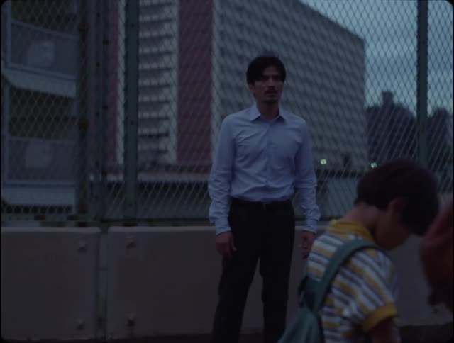 Boy walks by man standing near fence in front of building