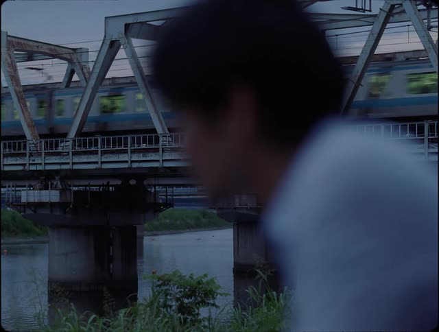 A black man sitting close to a river near a train bridge with a train passing by