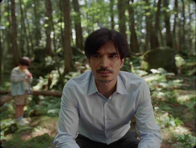 A man crouches down in a forest wearing a shirt in front of a girl