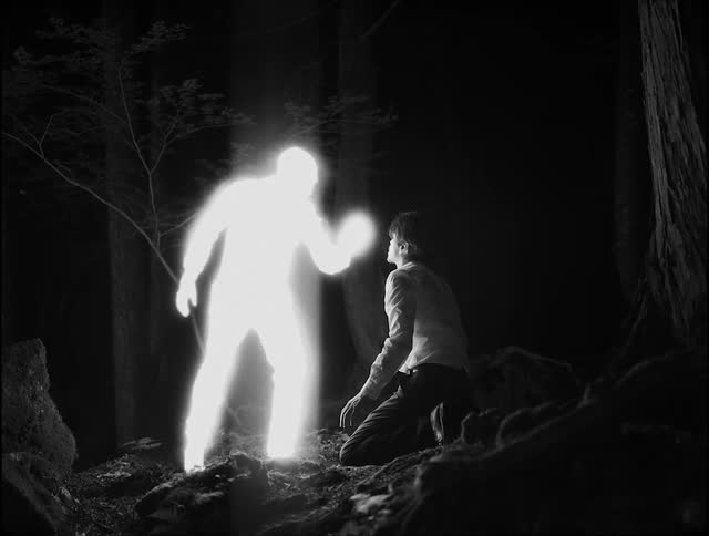A man is on his knees in the woods at night as a brightly glowing man touches him on the face and illuminates the surrounding area