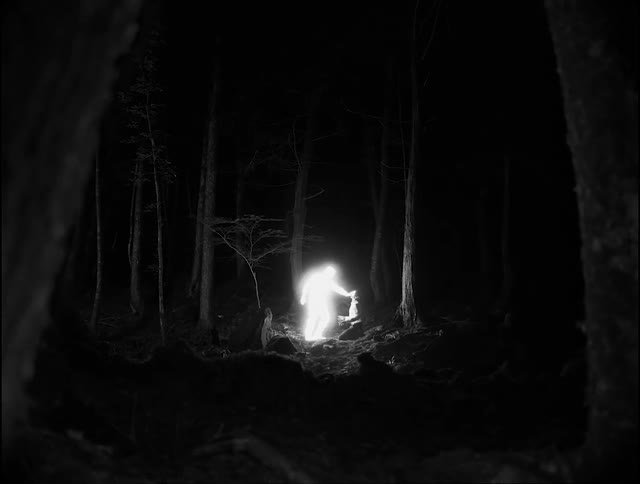 A man is on his knees in the woods at night as a glowing man touches him and illuminates the surrounding area