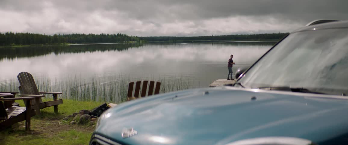 In the background a man on a deck is photographing a lake in the foreground on the right theres a car and on the left there are benches and a person passing by the car