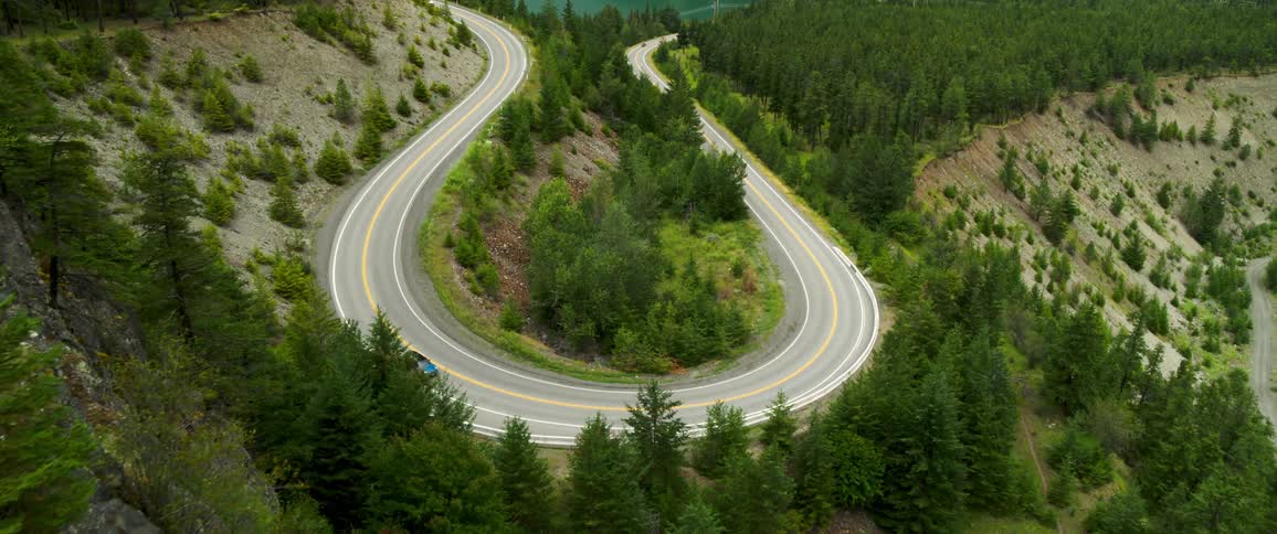 Aerial view of a circular road encircled by trees a car descending a mountain with a river in the background and an mountain