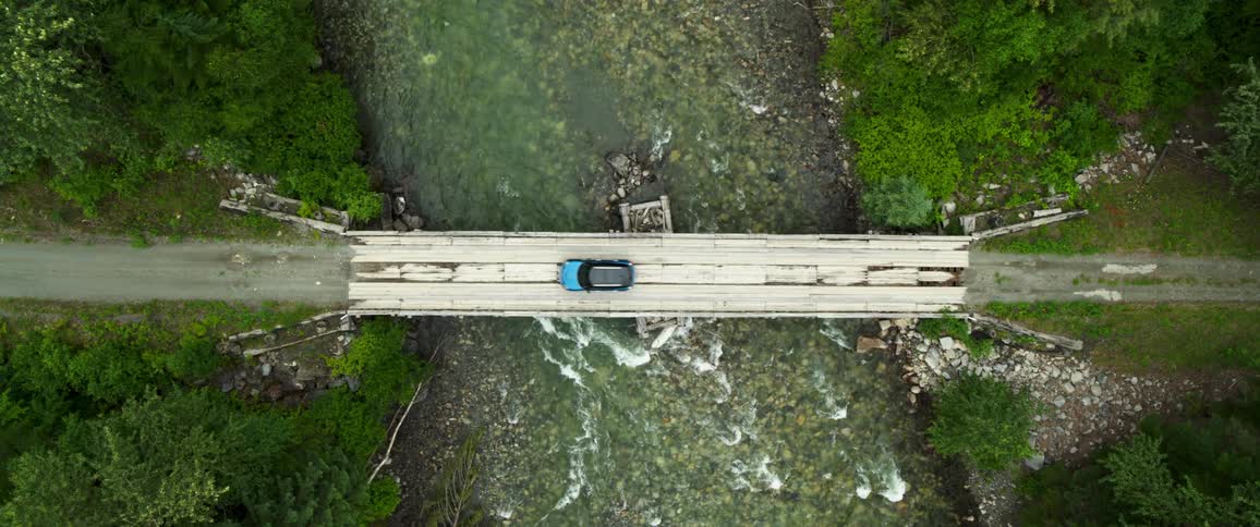 A blue car driving over a bridge over the river in the forest