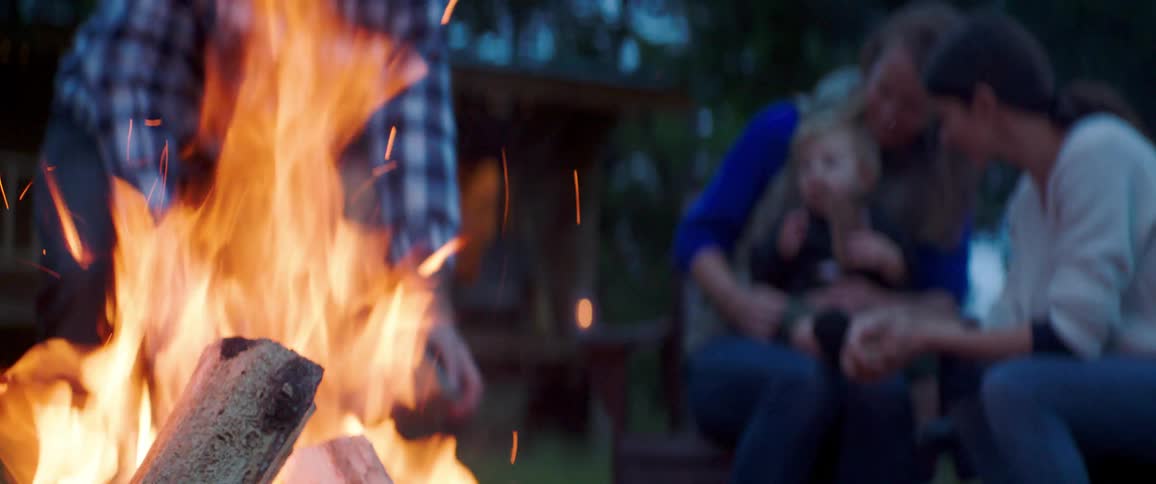 View of a campfire with a father putting firewood and sea holding his son next to a woman