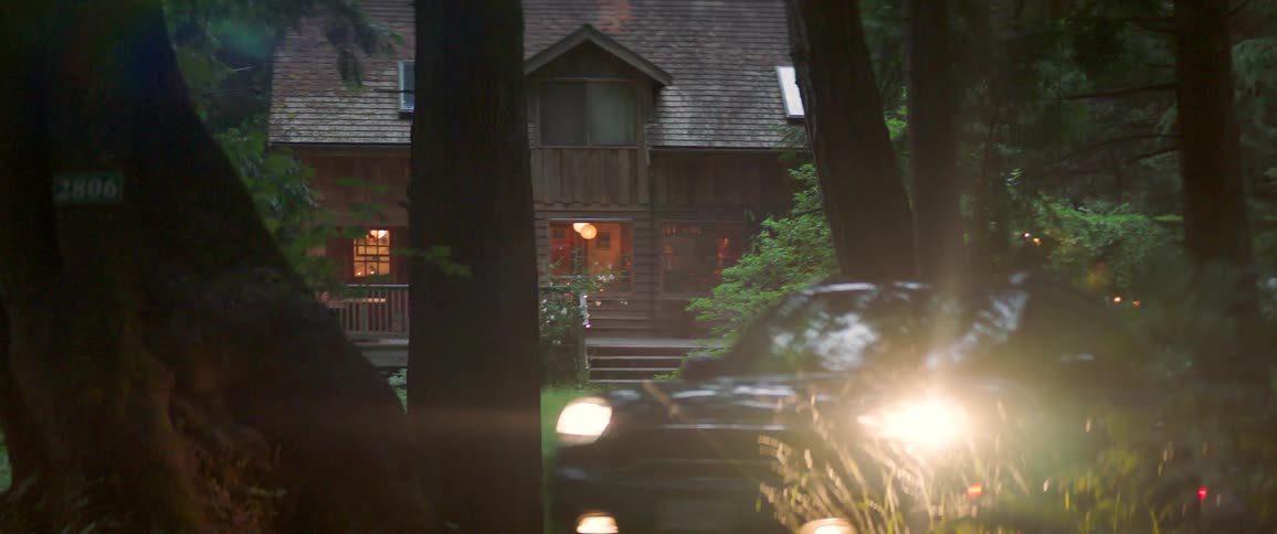 A car with headlights on drives away from a house in the woods