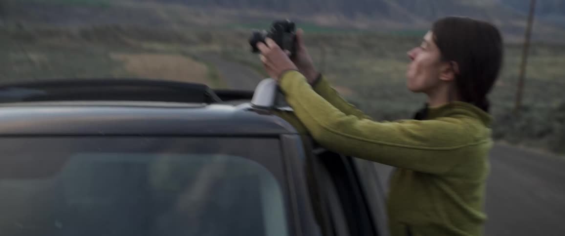 A lady sits on the window of a parked car taking pictures with a camera in the evening