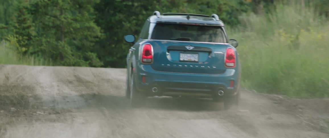 Blue mini cooper drives down gravel road through trees