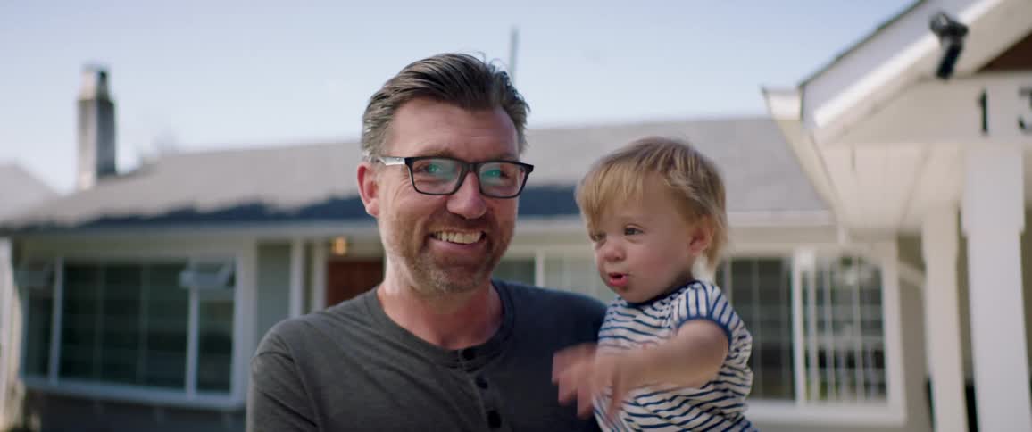 Father stands outside home holding son and smiling