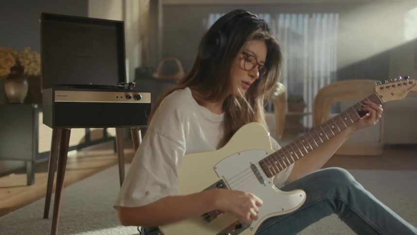 A woman with glasses and headphones plays a white guitar on a white rug with a vinyl player in the background