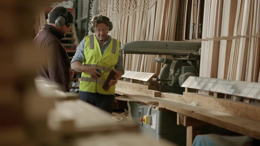 Man in a factory picking up a piece of wood measuring it and showing it to his colleague next to him