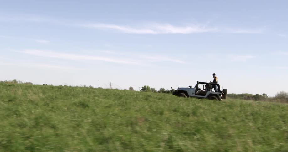 Two people stand in a jeep as it crosses a field