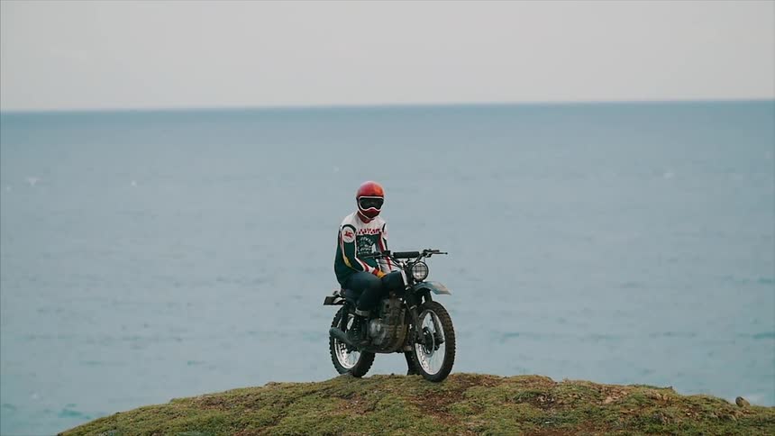 A person sitting on a motorbike on a hill in front of a body of water