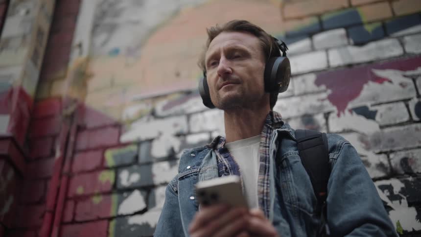 Man using smartphone listens to headphones while standing next to mural covered building