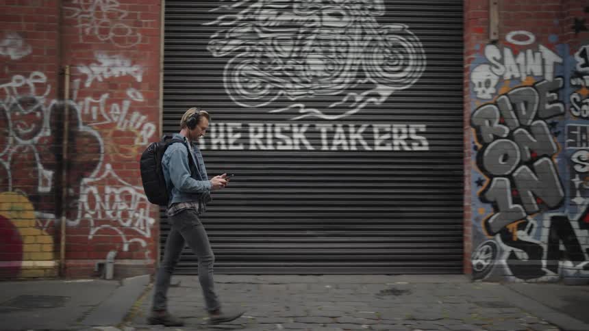 A man with headphones a black backpack a denim jacket and gray jeans walks down the street passing by buildings with graffiti while using a smartphone