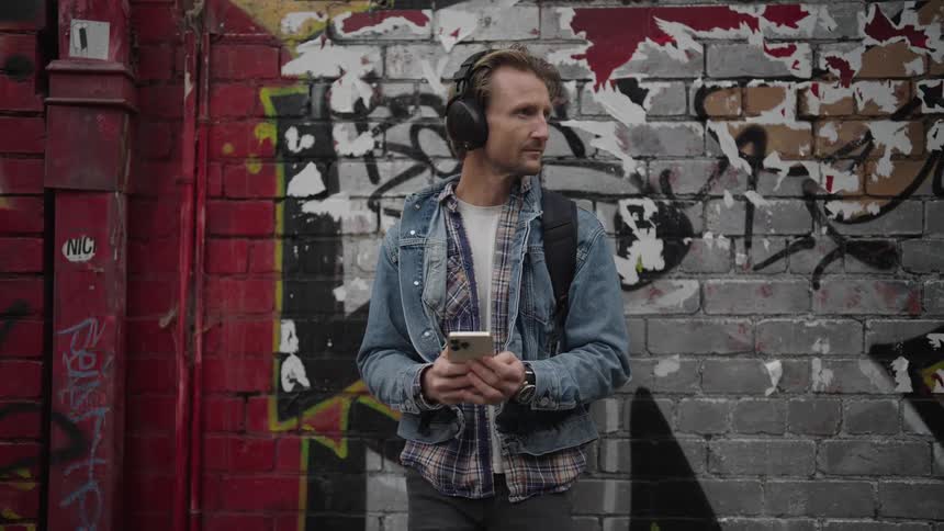 Man listens to music and uses smartphone while standing next to graffiti covered building