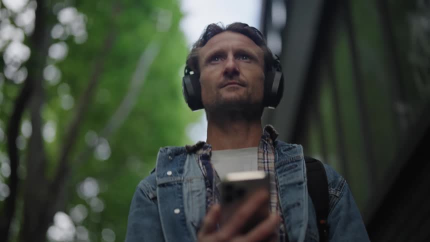 A man in a denim jacket walks down the street while using a smartphone and wearing headphones