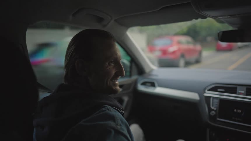 A blonde man wearing a denim jacket sits in the front seat of a car talking to someone and sharing a laugh