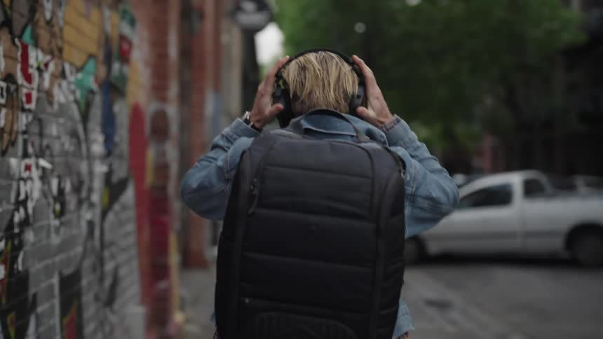 Man in a denim jacket walking around a left corner with a black backpack and putting on a headphone